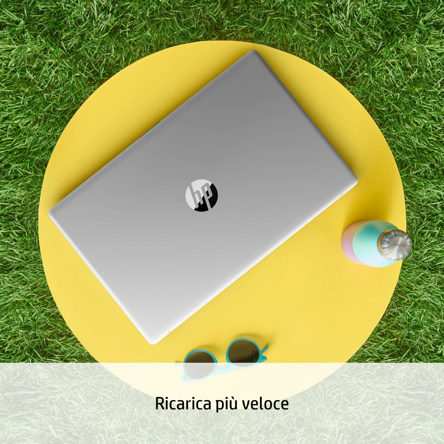 Un laptop argentato su un tavolo giallo sull'erba, con occhiali da sole e bottiglia vicini.
