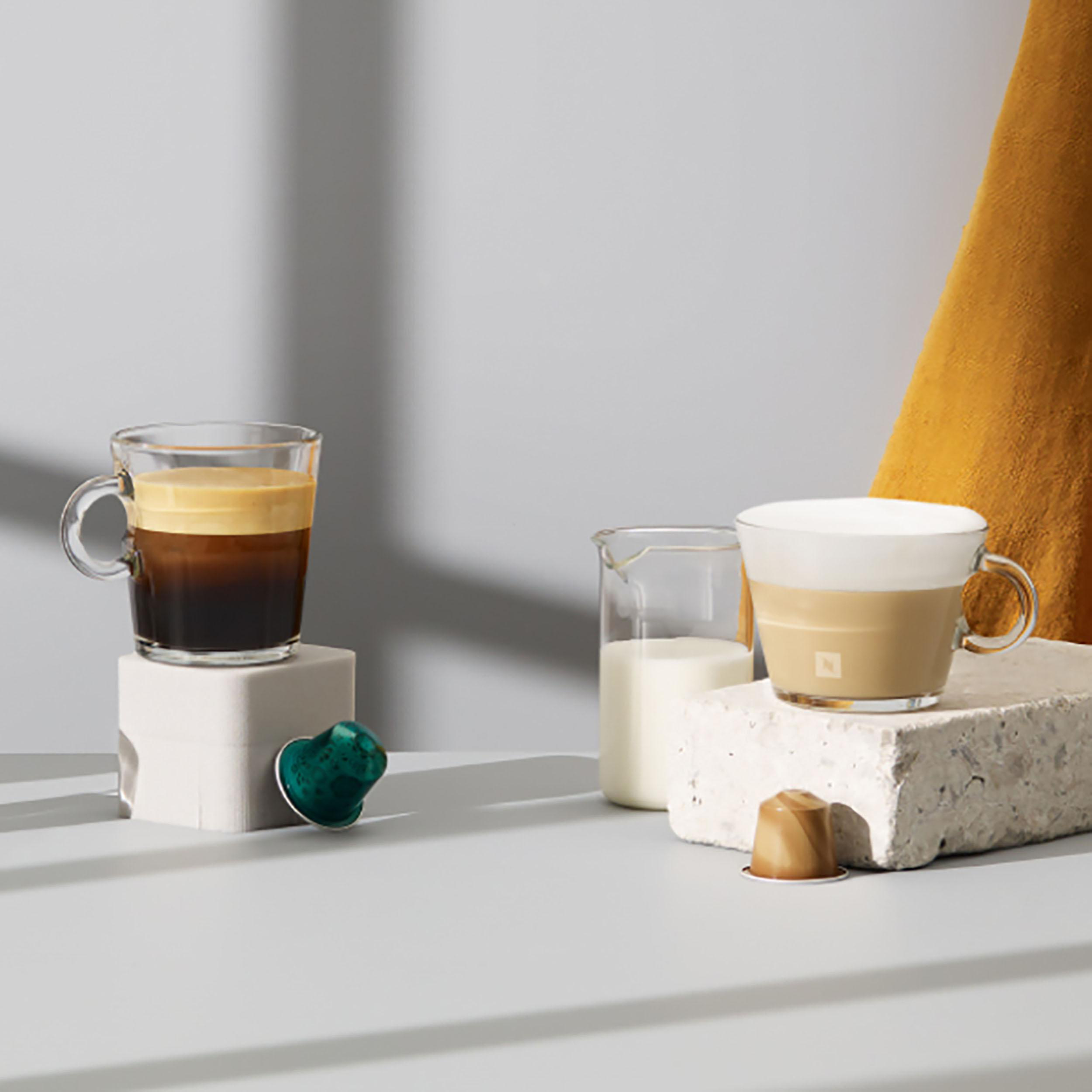 Zwei Kaffeetassen, Milch in einem Becher, Nespresso-Kapseln auf Steinen.