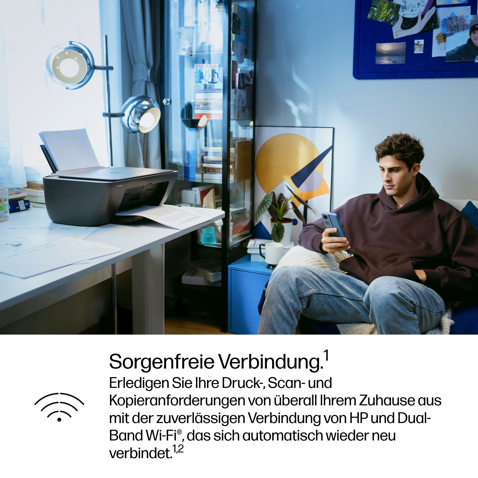 Ein Mann sitzt auf einem Sofa und schaut auf sein Handy. Ein Drucker steht auf einem Schreibtisch.