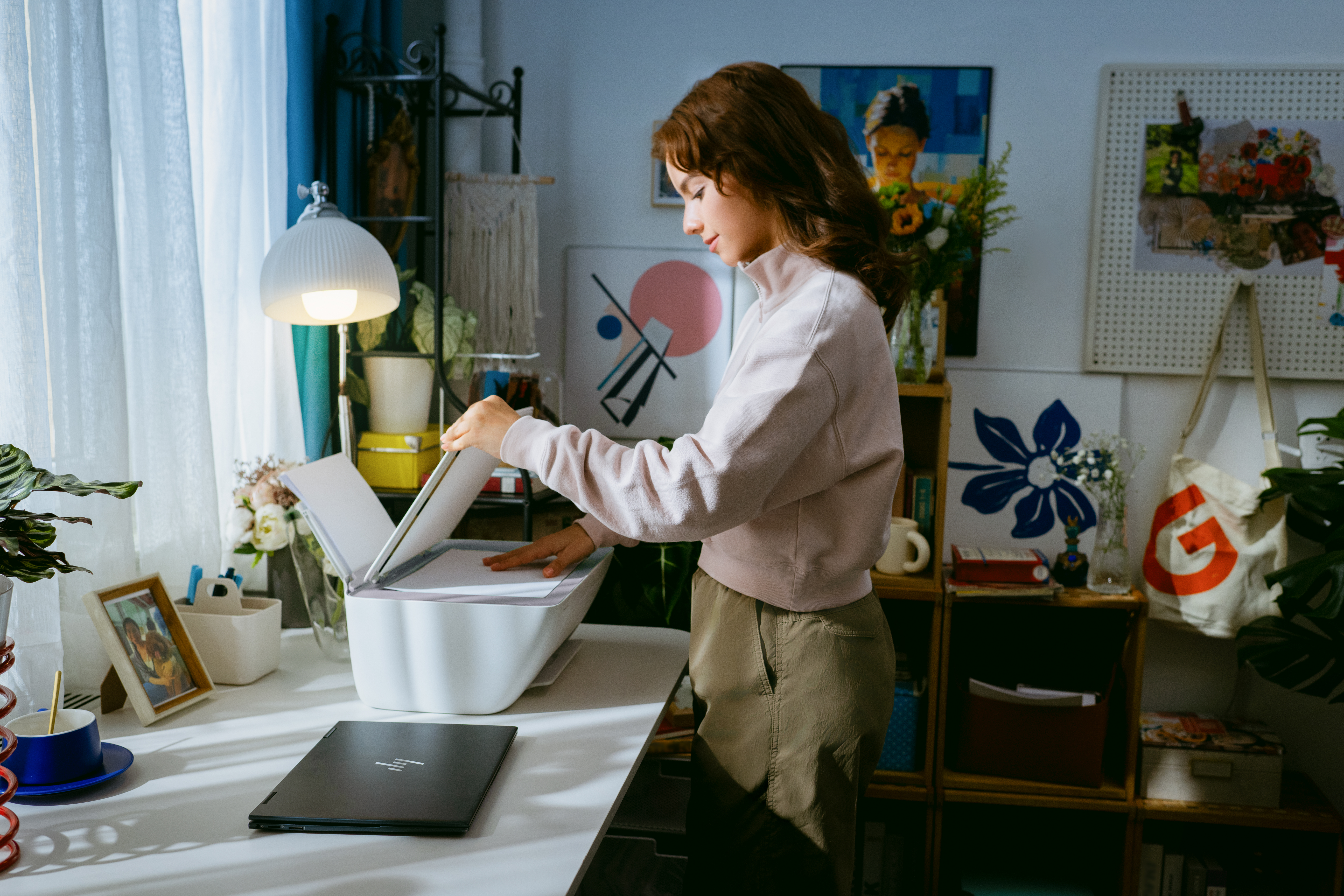 Egy nő rózsaszín pulóverben fehér nyomtatót használ egy laptopos asztalon.