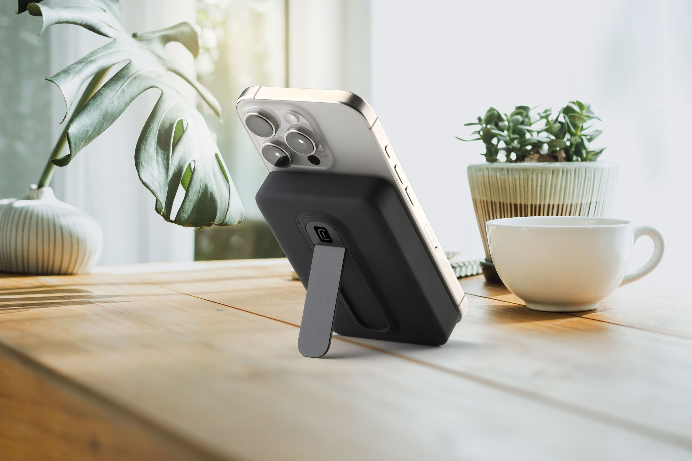 Smartphone plateado con soporte negro en mesa de madera junto a planta y taza.