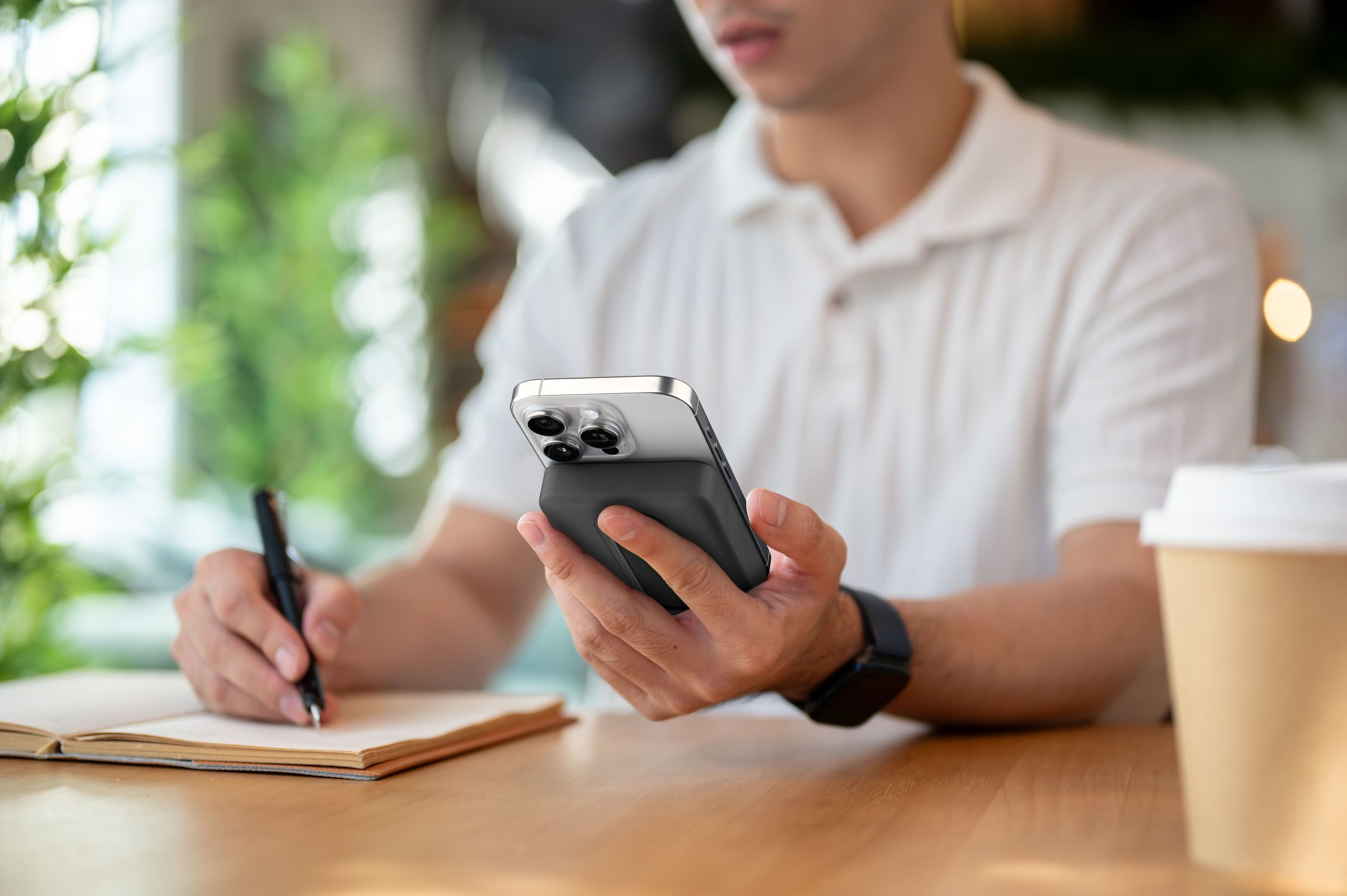 Un hombre escribe en un cuaderno mientras sostiene un teléfono con cargador. Taza de café cerca.