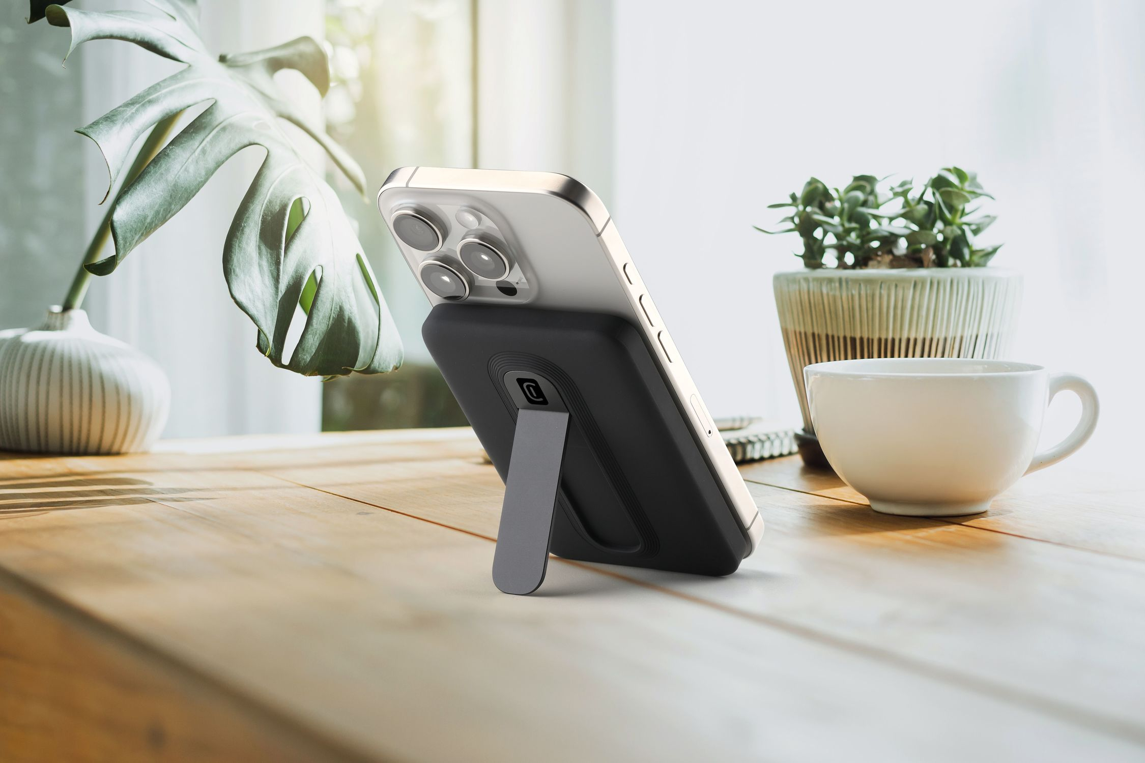 Un smartphone con soporte en mesa de madera, junto a planta y taza.