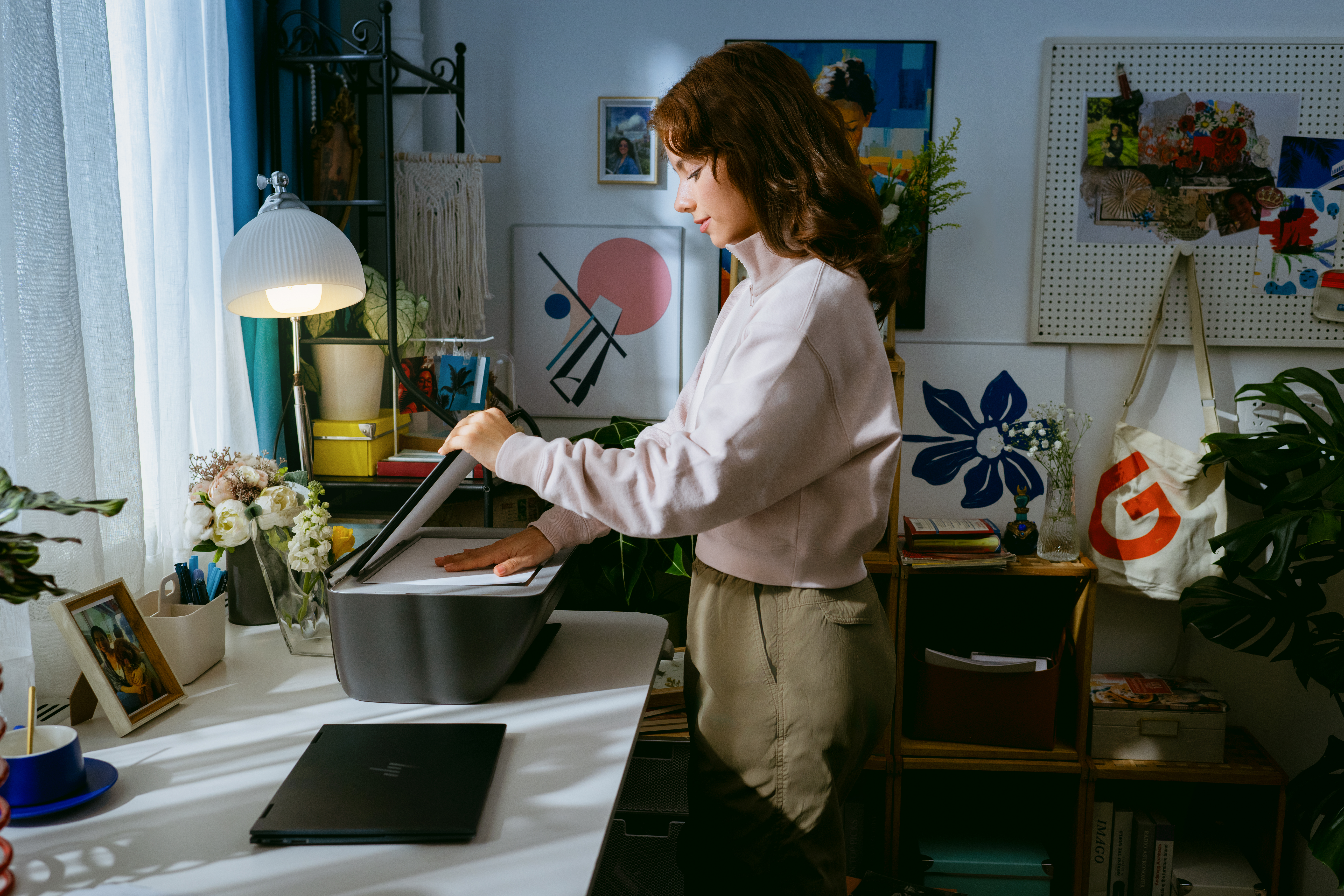 Eine Frau mit braunen Haaren benutzt einen Drucker an einem Schreibtisch mit Laptop und Lampe.