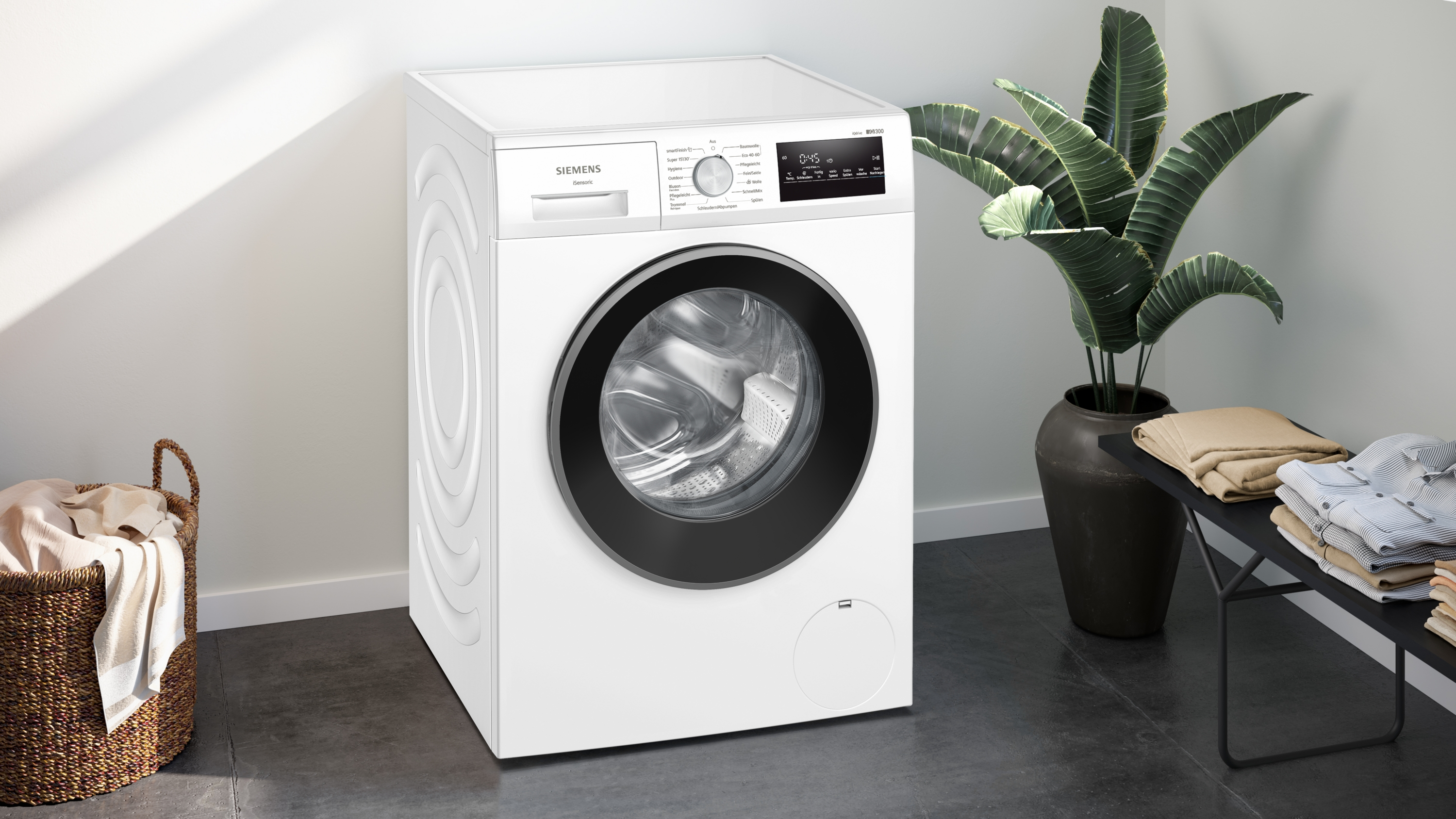Eine weiße Siemens Waschmaschine mit schwarzer Tür steht neben einem Korb und einem Regal mit Kleidung.