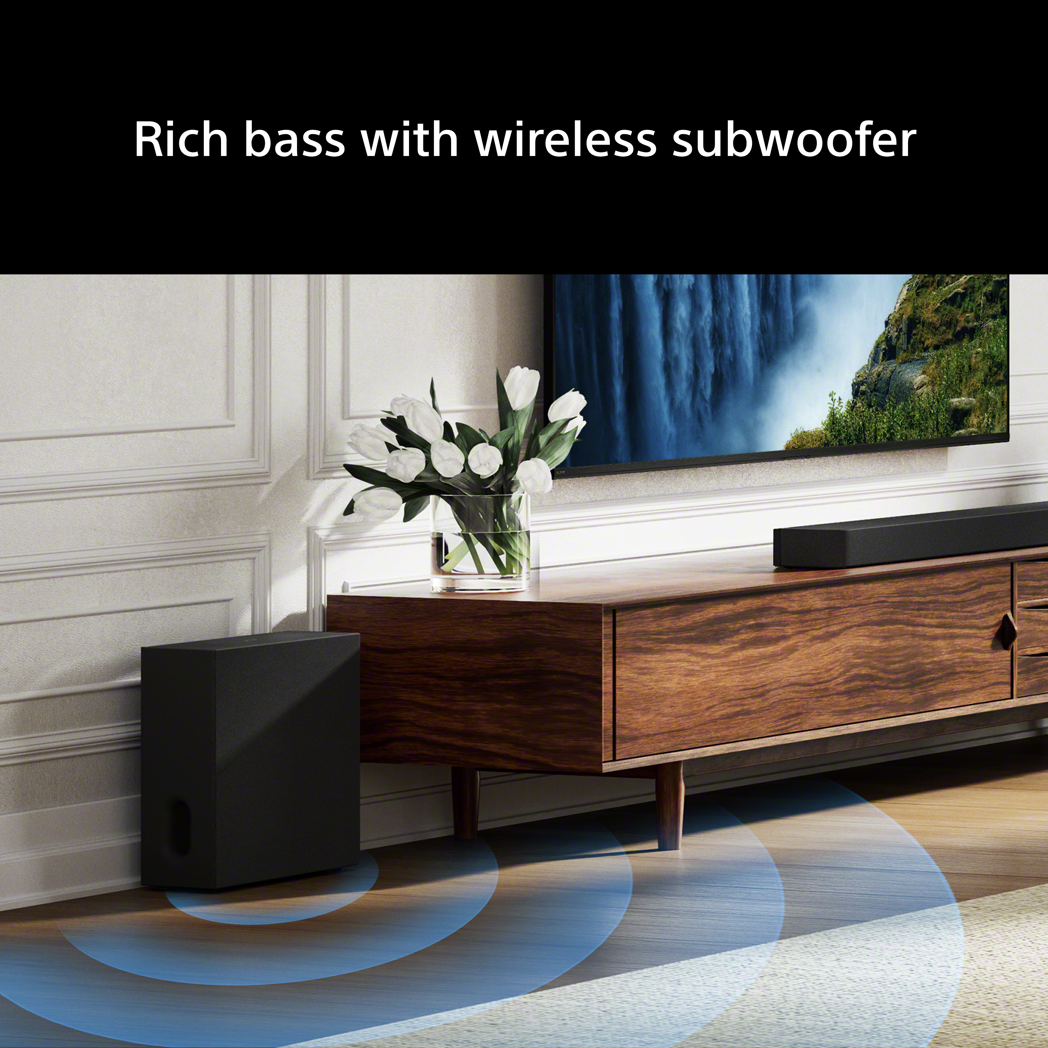 Un subwoofer nero è su un pavimento in legno. Una soundbar poggia su un mobile in legno.