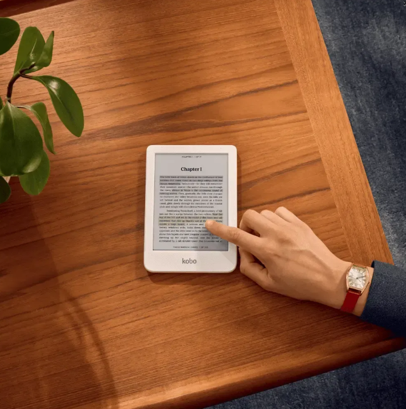 Dłoń dotyka czytnika e-booków na drewnianym stole obok rośliny.