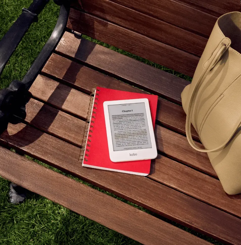 Biały czytnik e-booków i czerwony notatnik leżą na drewnianej ławce na zewnątrz.