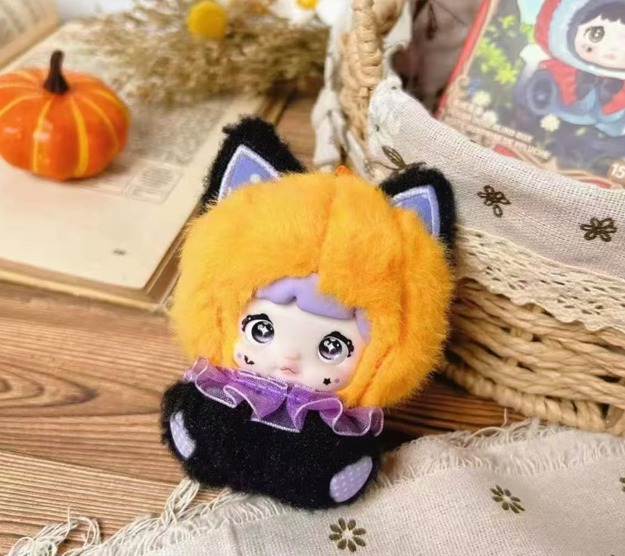 Eine Kuschelpuppe mit orangefarbenem Haar und Katzenohren sitzt neben einem Buch und einem Kürbis.