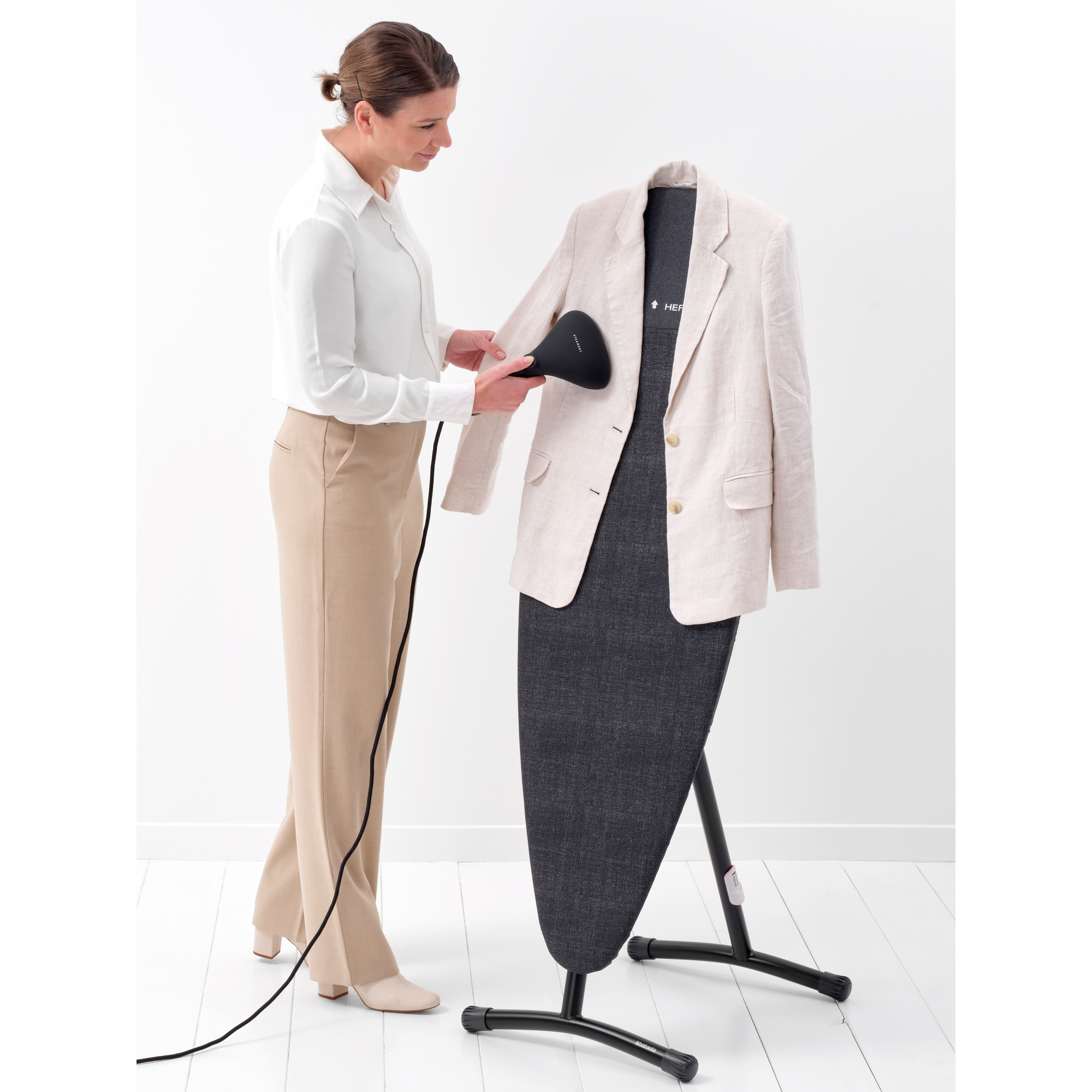 Frau bügelt eine beige Jacke mit einem Handdampfer vor einem Bügelbrett.