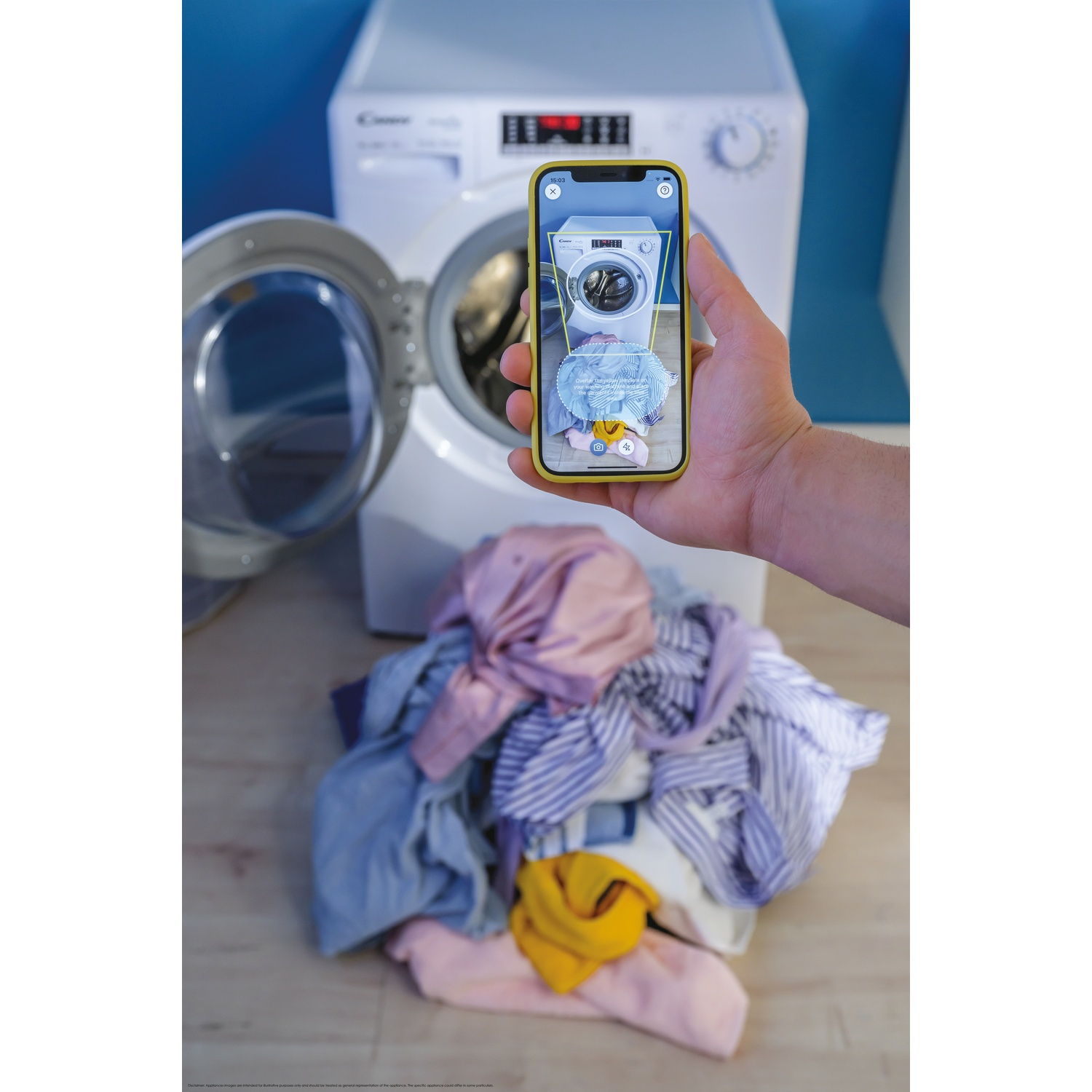 Una mano tiene uno smartphone che mostra una lavatrice con biancheria. Una vera lavatrice con porta aperta è vicina.