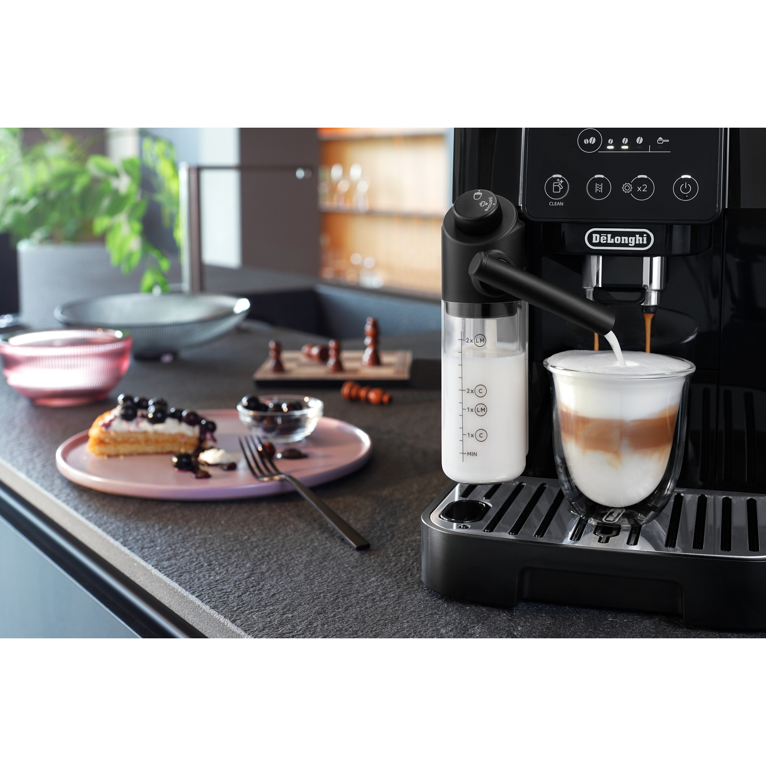 A black coffee machine brews a latte into a glass. Next to it, a cake with blueberries.
