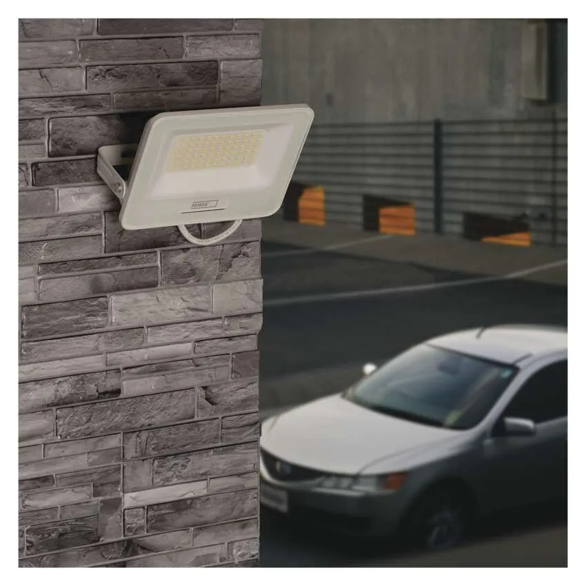 Egy fehér LED-es reflektor egy szürke téglfalra van szerelve. Egy ezüst színű autó parkol a közelben.