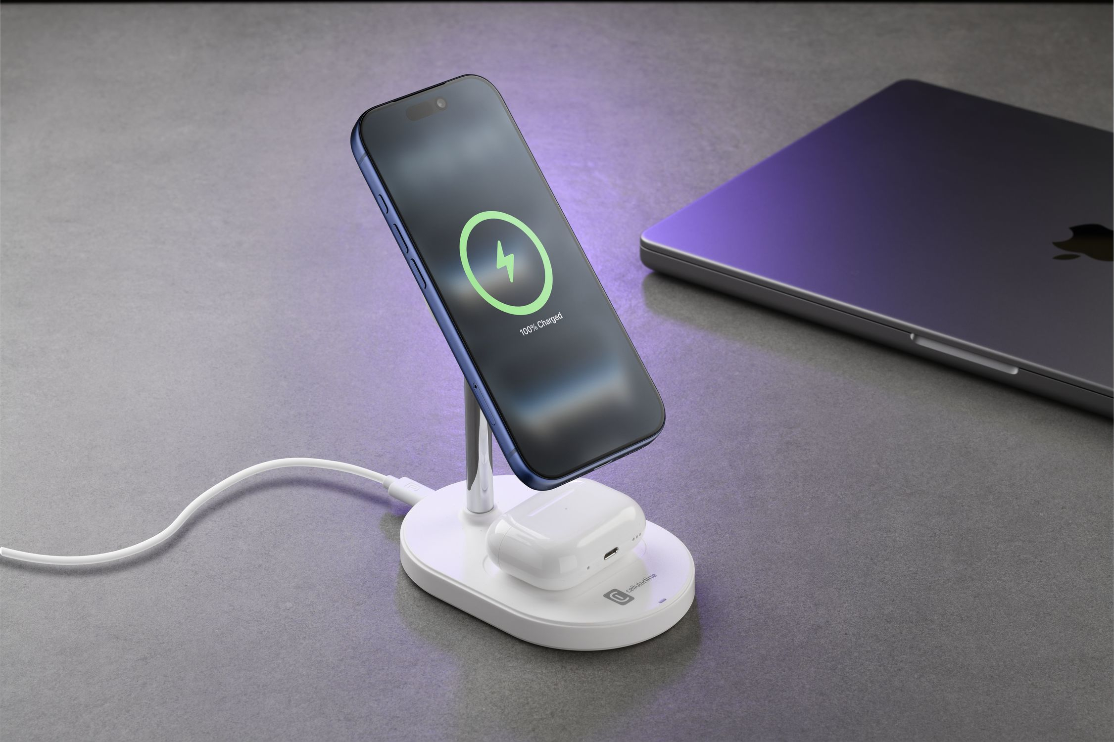 Un iPhone azul en un soporte cargador con auriculares y portátil cerca.