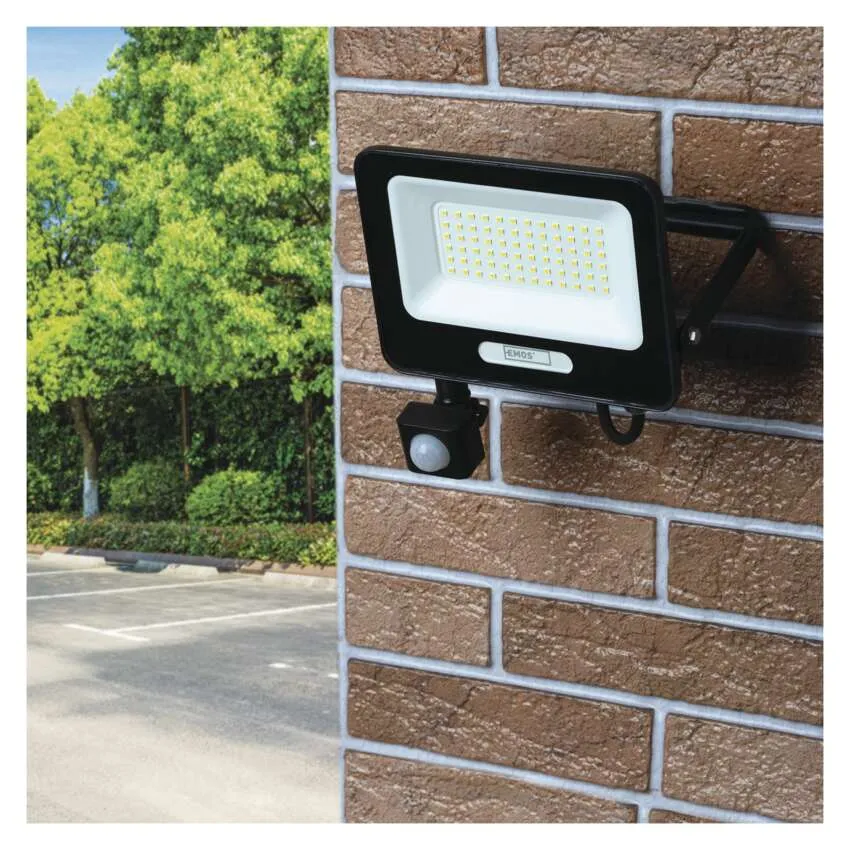 Fekete LED-reflektor mozgásérzékelővel téglakerítésen, zöld fák és parkoló a háttérben.