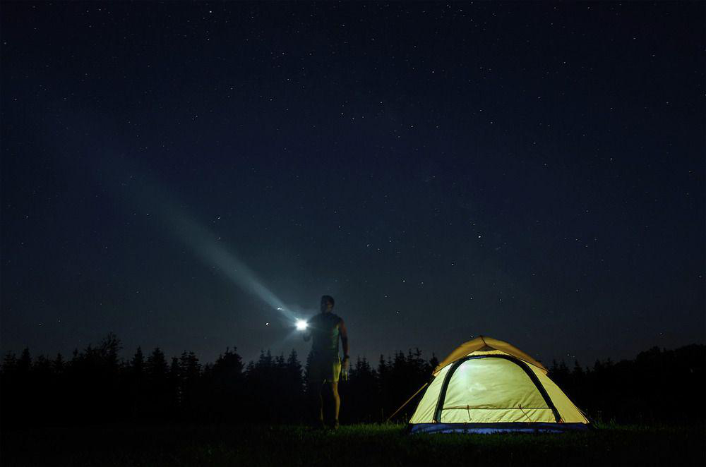 Ein Mann mit einer Taschenlampe neben einem beleuchteten Zelt unter einem sternenklaren Nachthimmel.