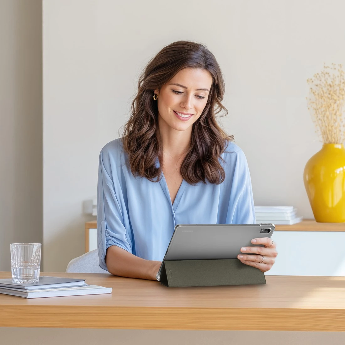 Frau in blauem Hemd benutzt Tablet am Holztisch mit Glas Wasser und Büchern.