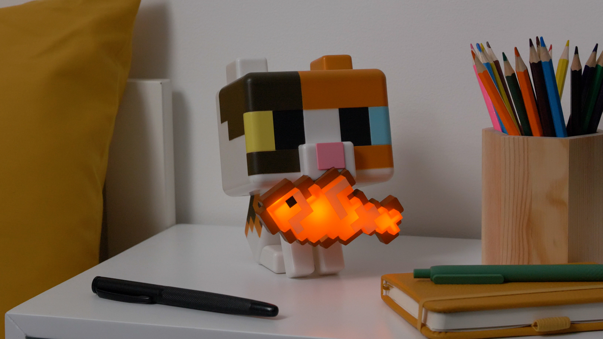 Eine pixelige Katzenlampe mit einem orangefarbenen Fisch im Maul steht auf einem Schreibtisch.