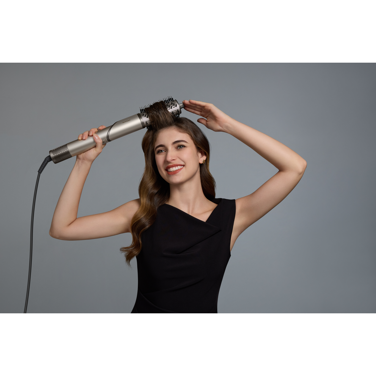 Een vrouw met bruin haar stylet haar haar met een krultang tegen een grijze achtergrond.
