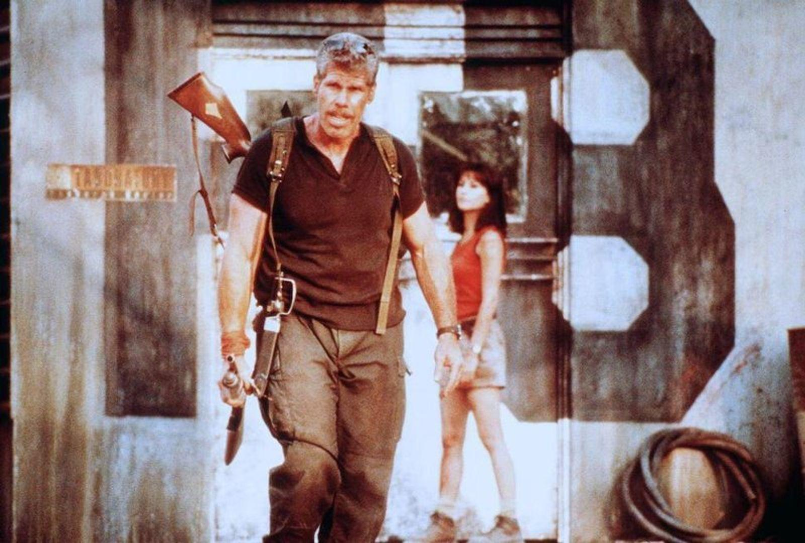 Ein Mann mit Gewehr trägt eine Frau mit rotem Top und kurzem Rock, die ihn begleitet.