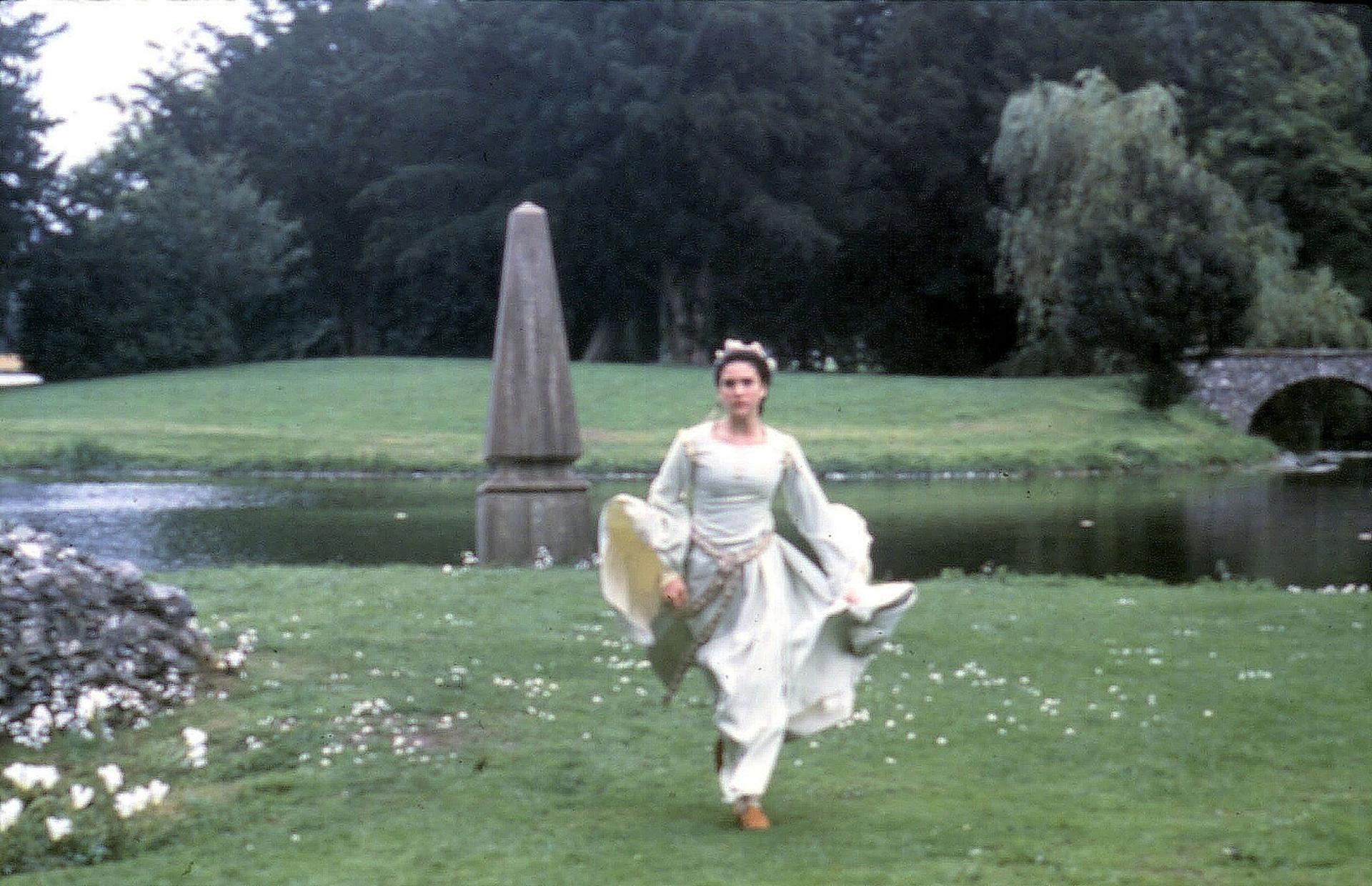 Eine Frau in einem weißen Kleid läuft auf einer grünen Wiese mit einem Obelisken und Bäumen.