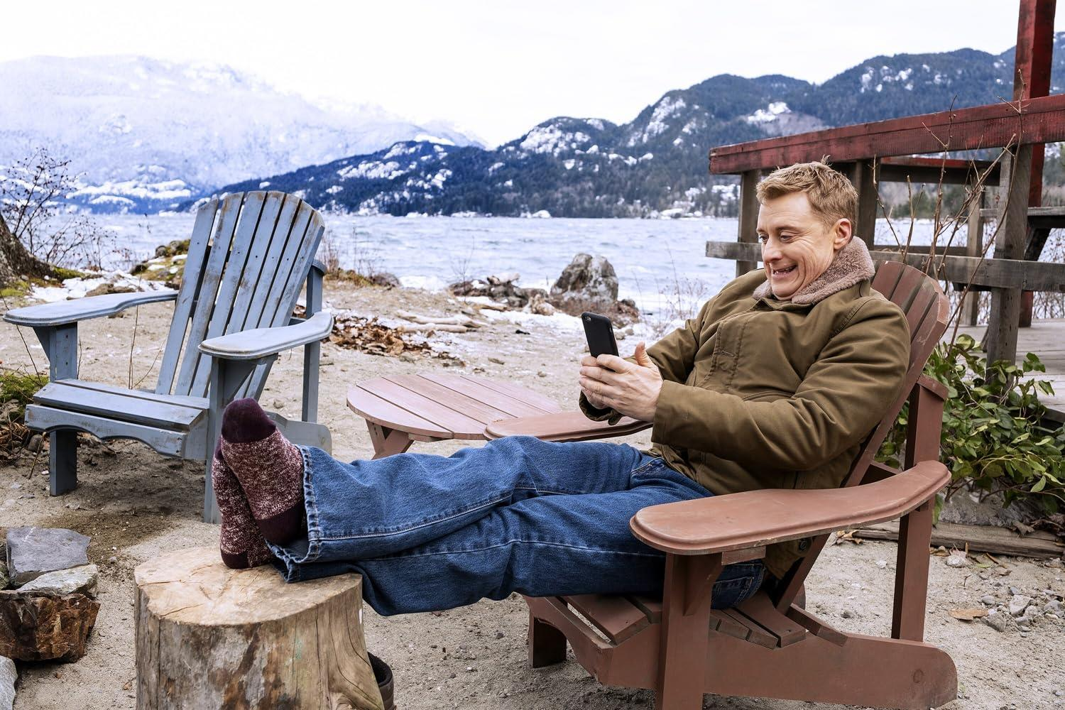 Ein Mann sitzt draußen auf einem Stuhl und schaut auf sein Handy. Berge im Hintergrund.