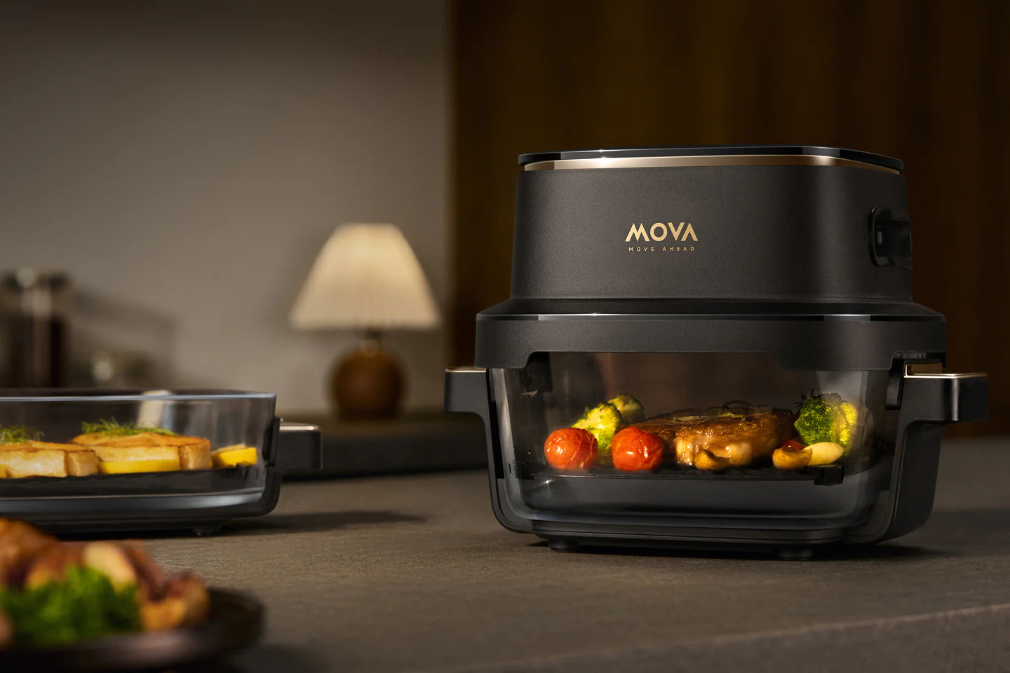 Czarny piekarnik MOVA ze stekiem, pomidorami, brokułami; obok szklana tacka z tostami.