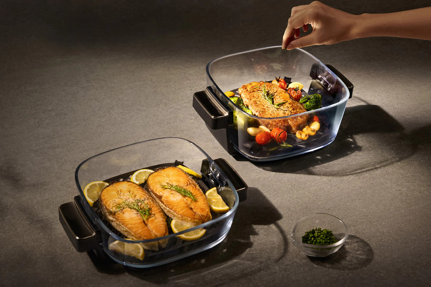 Dwie szklane potrawy z pieczonym łososiem i warzywami, ręka posypuje ziołami.