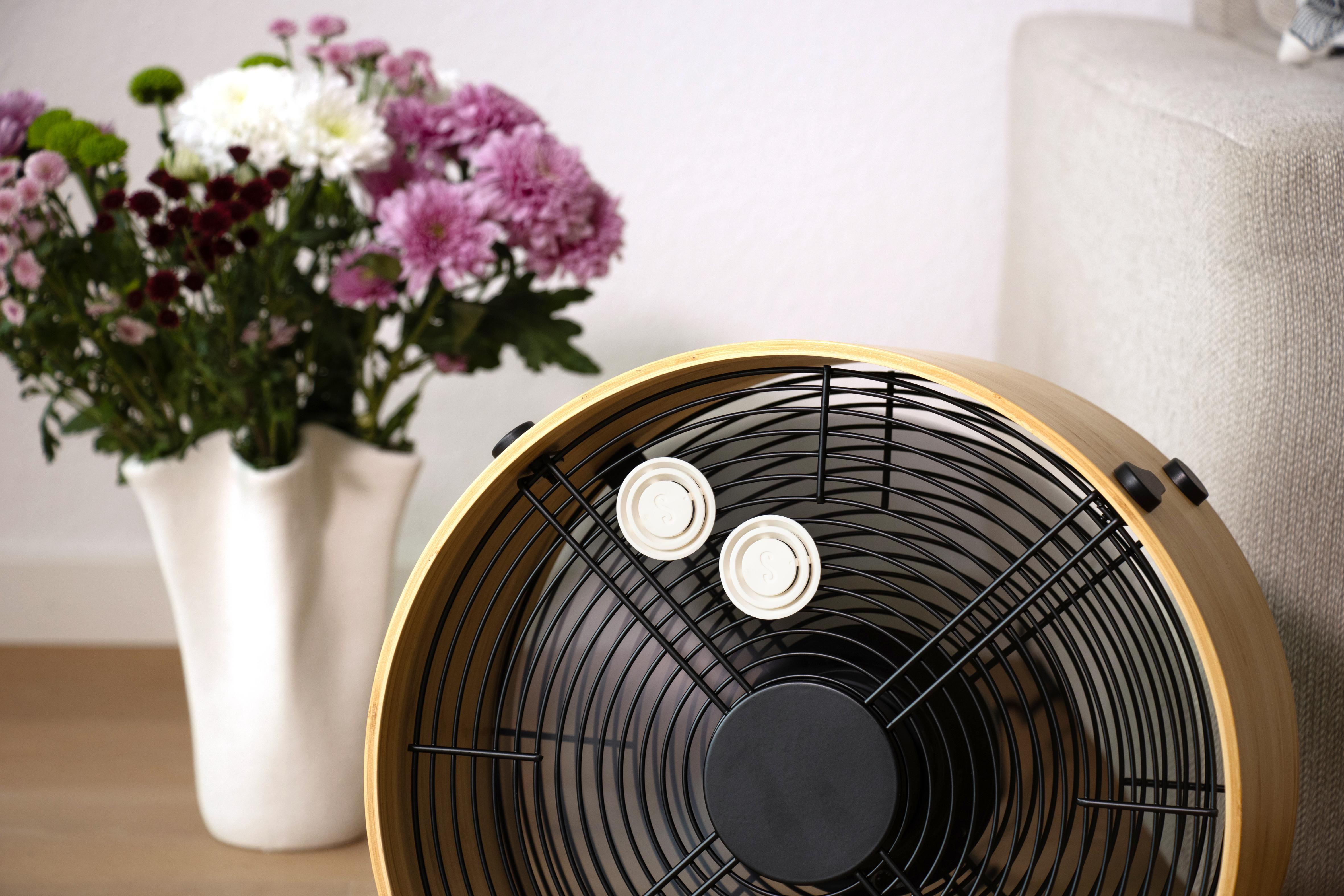 Ein Ventilator mit Holzrahmen steht neben einer Vase mit bunten Blumen.
