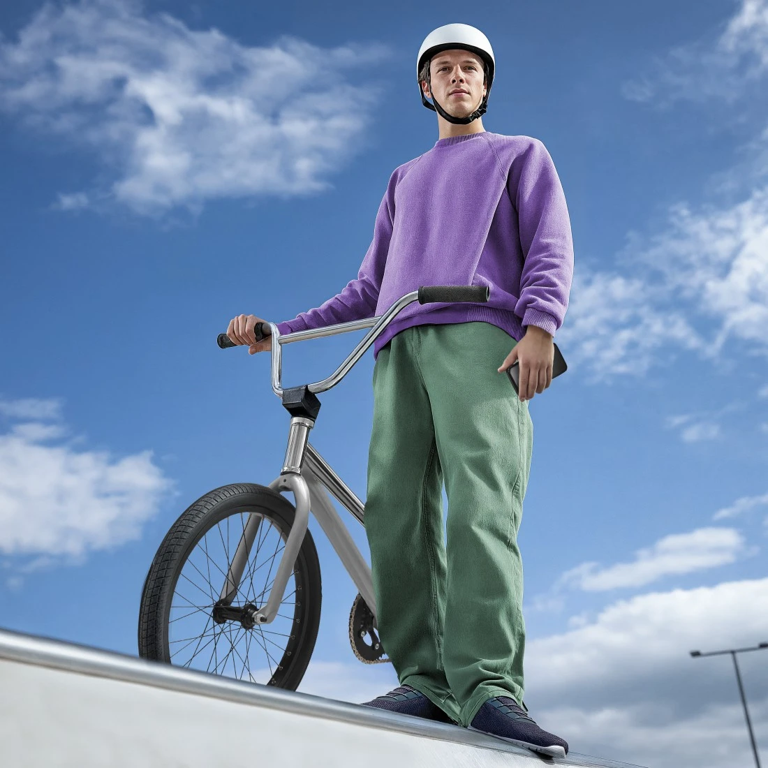 Ein Mann in lila Pullover und grüner Hose steht mit einem BMX-Rad bei klarem Himmel.