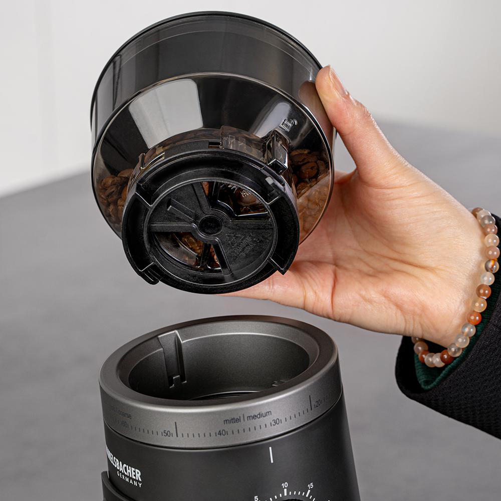 Hand hält Behälter mit Kaffeebohnen über Kaffeemühle mit Mahlgrad-Einstellung.
