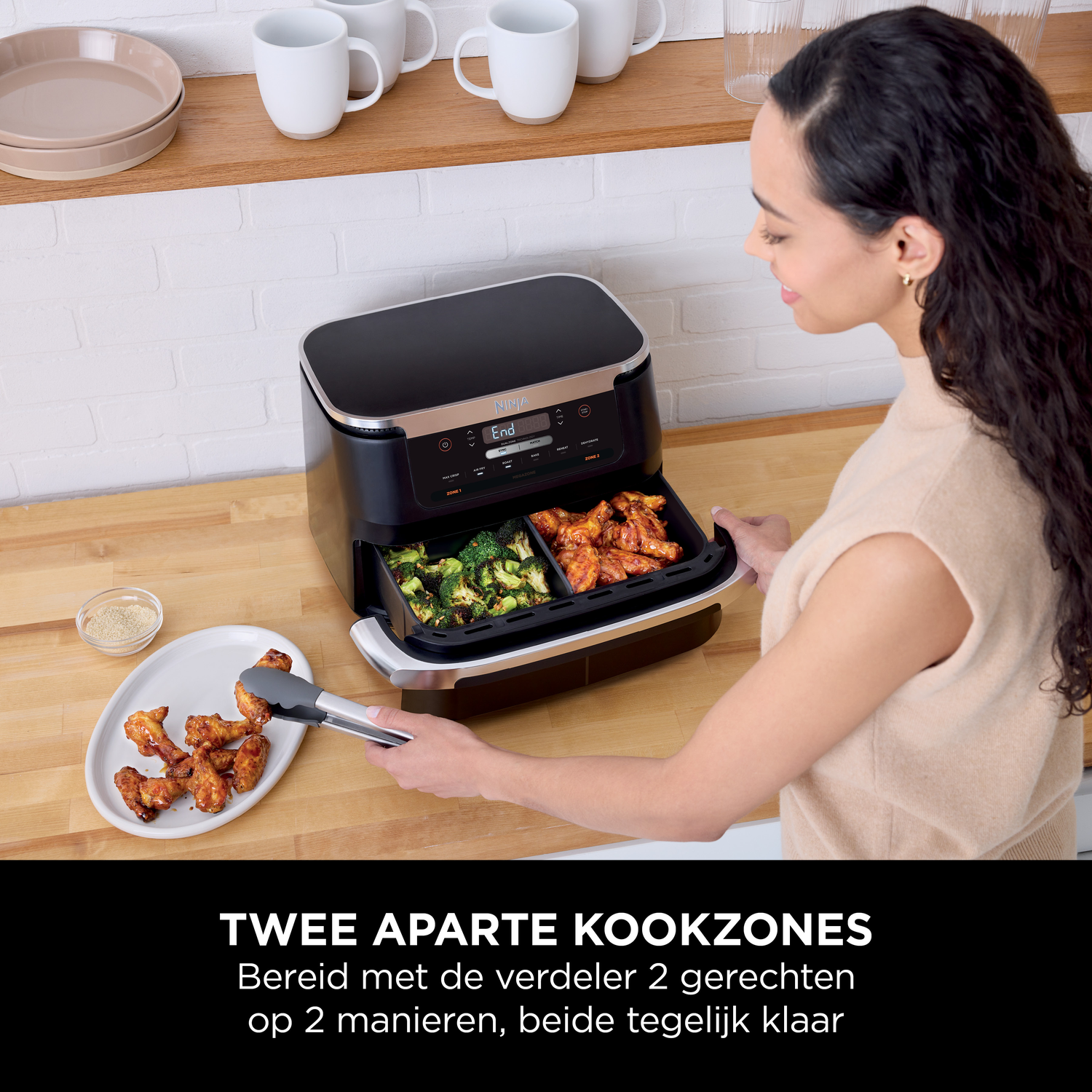Een vrouw haalt kippenvleugels uit een zwarte airfryer-mand met twee vakken.