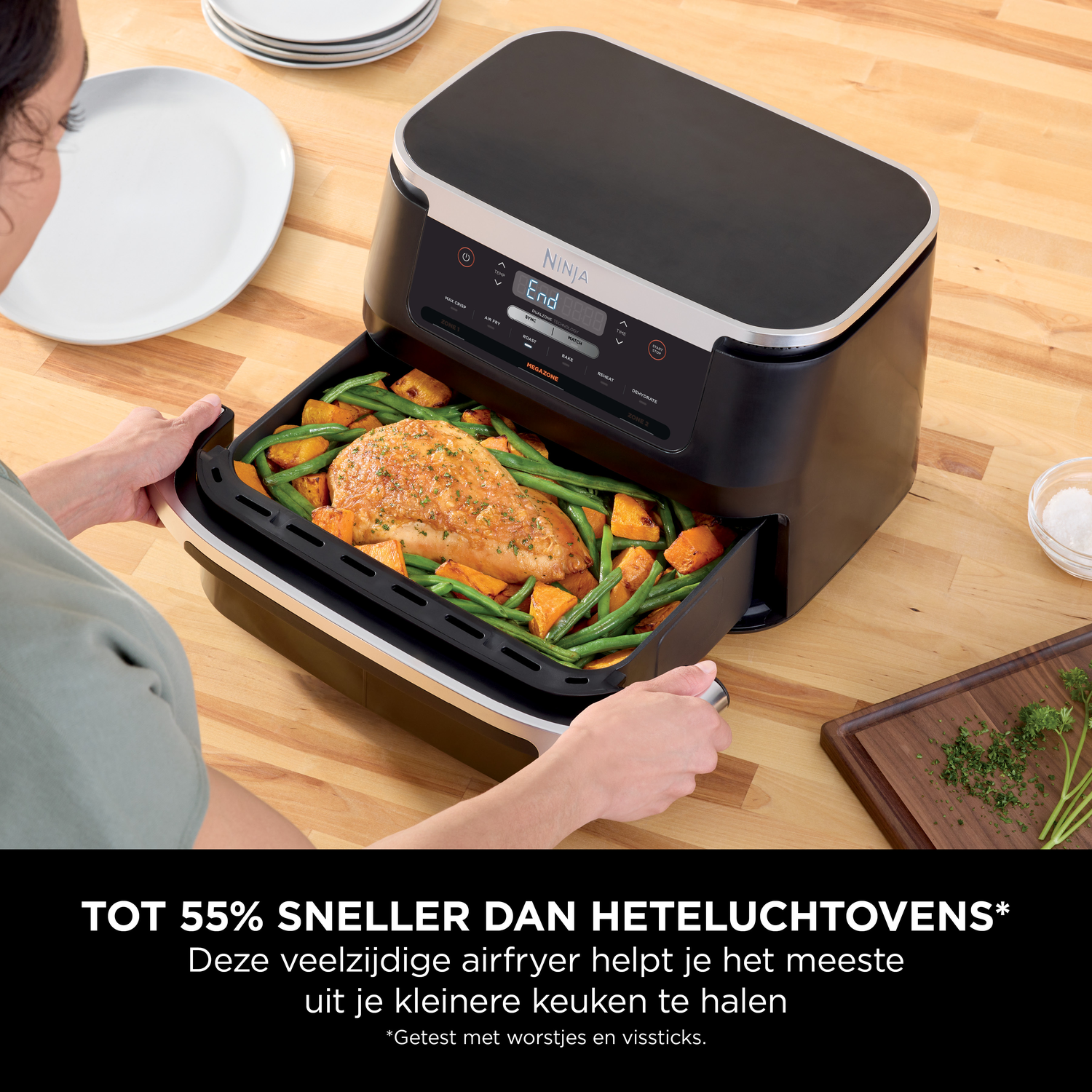Een vrouw trekt een airfryer mand met geroosterde kip en groenten eruit.