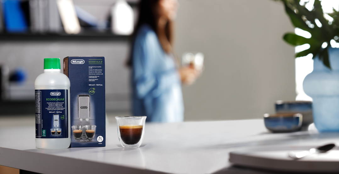 Eine Flasche und Schachtel DeLonghi Entkalker stehen auf einer Theke mit Espresso und verschwommener Person.