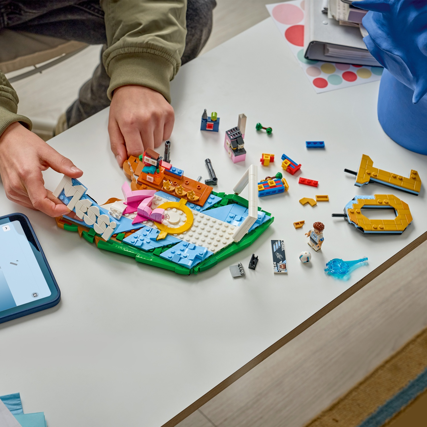 Mani che costruiscono LEGO colorati su un tavolo bianco. Un tablet mostra le istruzioni.