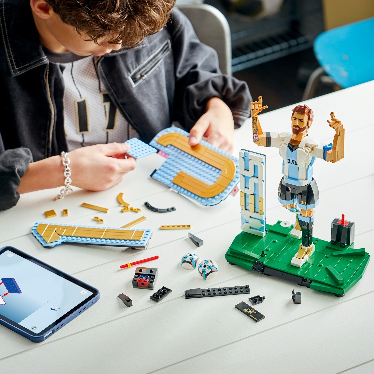 Un ragazzo costruisce un giocatore di calcio Lego con guida tablet, pezzi su tavolo bianco.