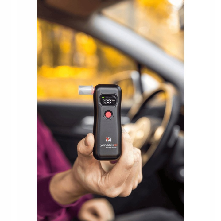 Dłoń trzyma czarny alkomat z ustnikiem i wyświetlaczem w samochodzie.