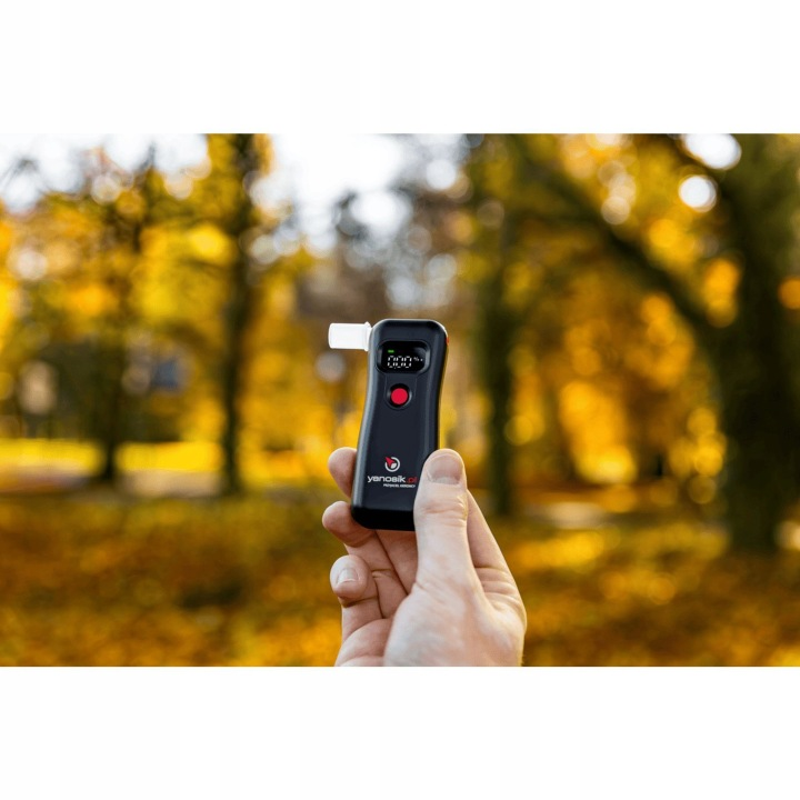 Ręka trzyma cyfrowy alkomat z ustnikiem, na zewnątrz jesienią.