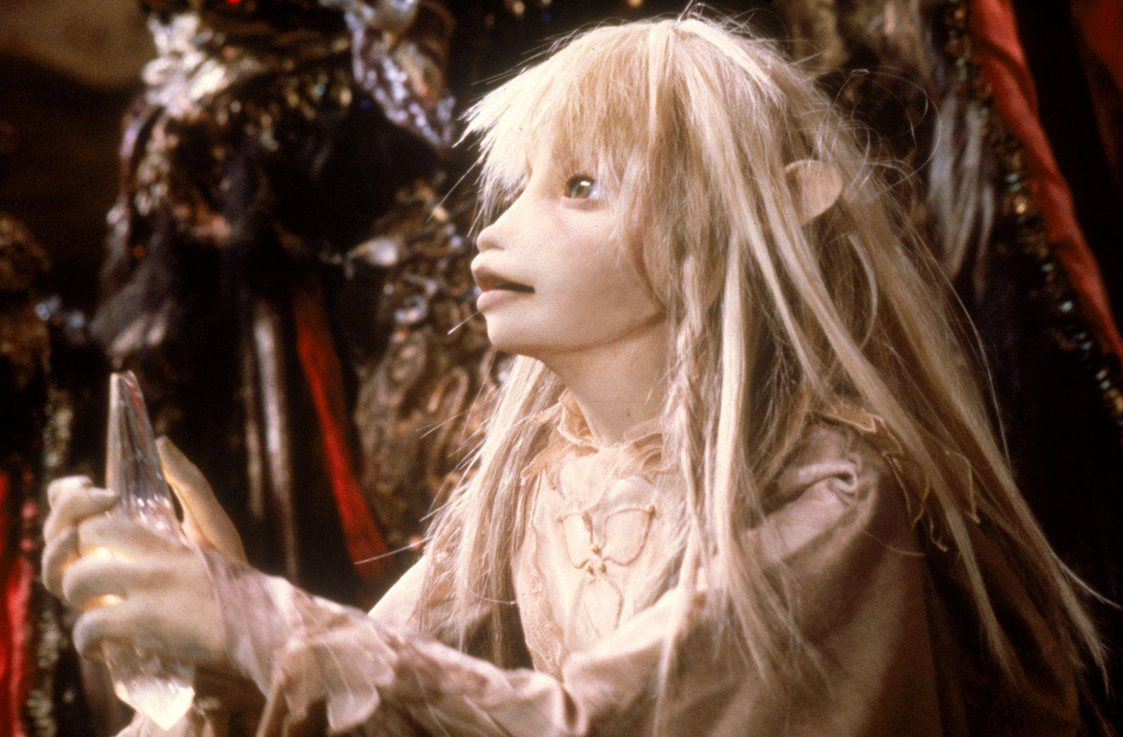 Ein hellhäutiges Wesen mit langen blonden Haaren hält einen Kristall.