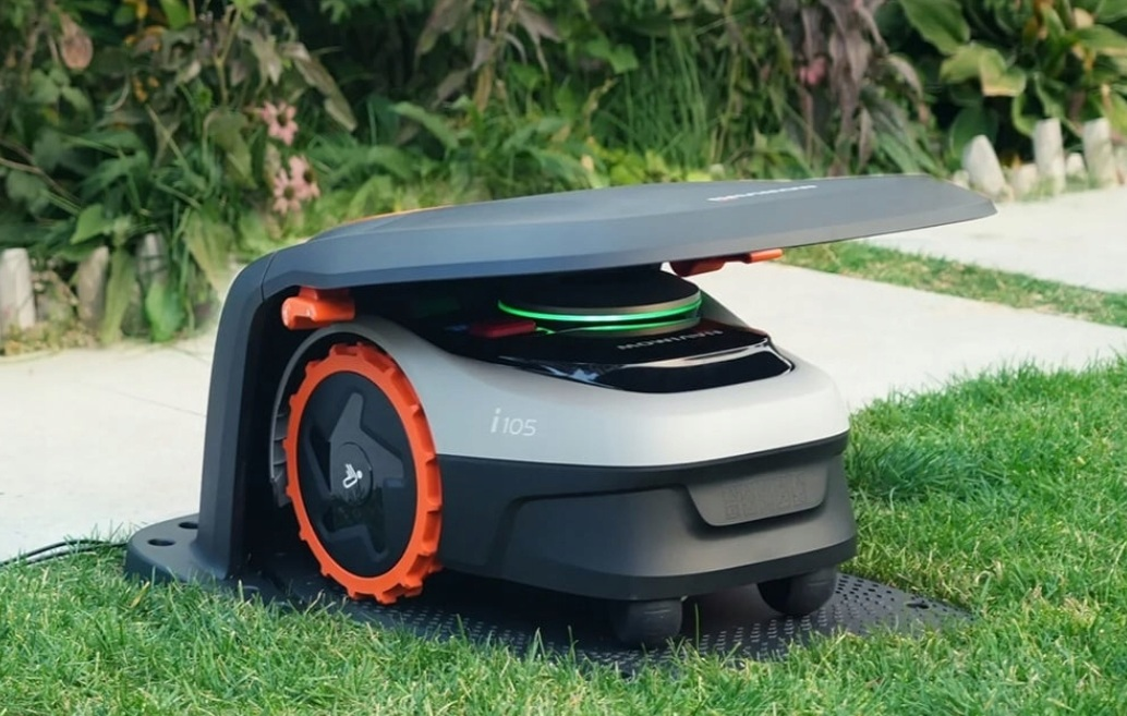 Robot koszący z pomarańczowymi kołami stoi na zielonym trawniku.