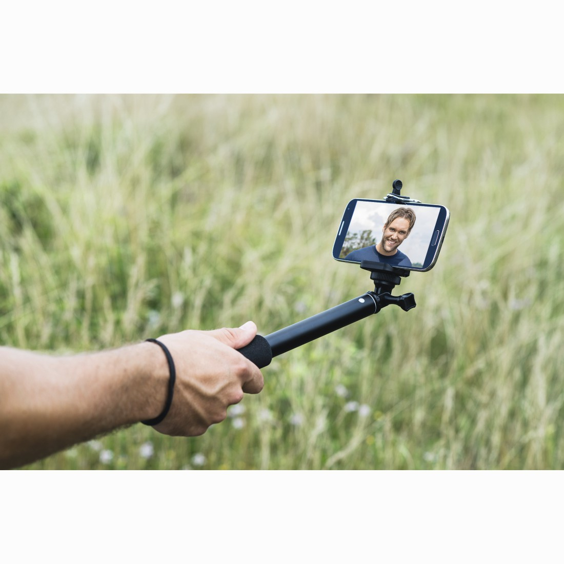 Bir adam dışarıda, arkasında çimen olan bir selfie çubuğuyla akıllı telefon tutuyor.