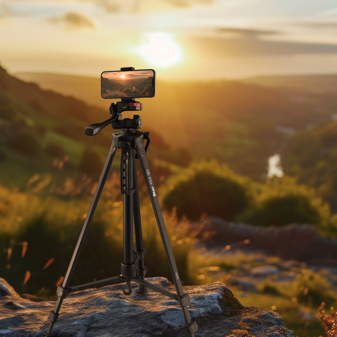 Kayaya yerleştirilmiş telefonlu siyah bir tripod, tepelik bir manzara üzerinde gün batımını yakalıyor.