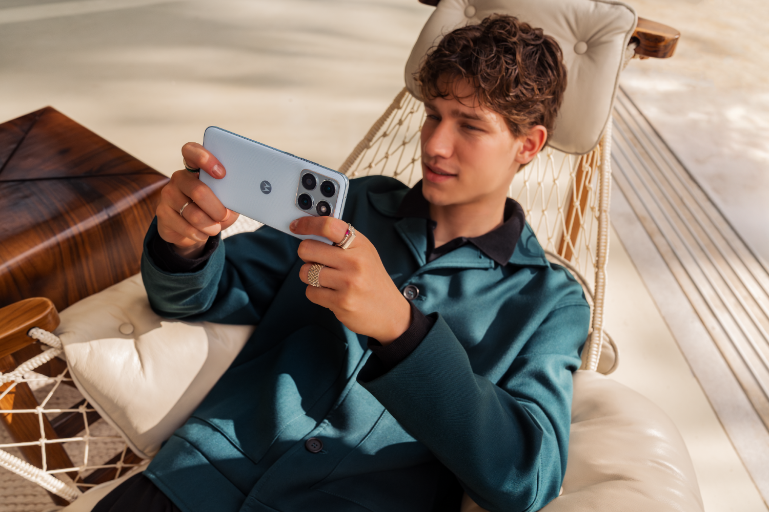 Młody mężczyzna w turkusowej kurtce trzyma jasnoniebieski telefon, siedzi w hamaku.