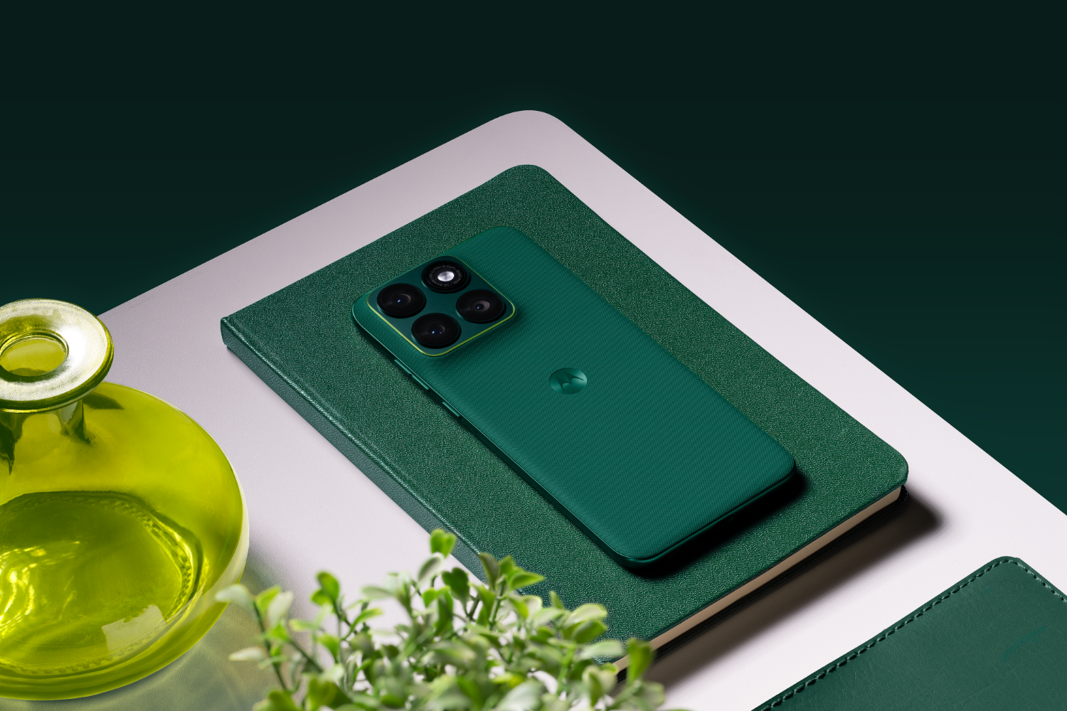 Zielony telefon leży na zielonym notatniku. W pobliżu znajduje się żółta waza i roślina.