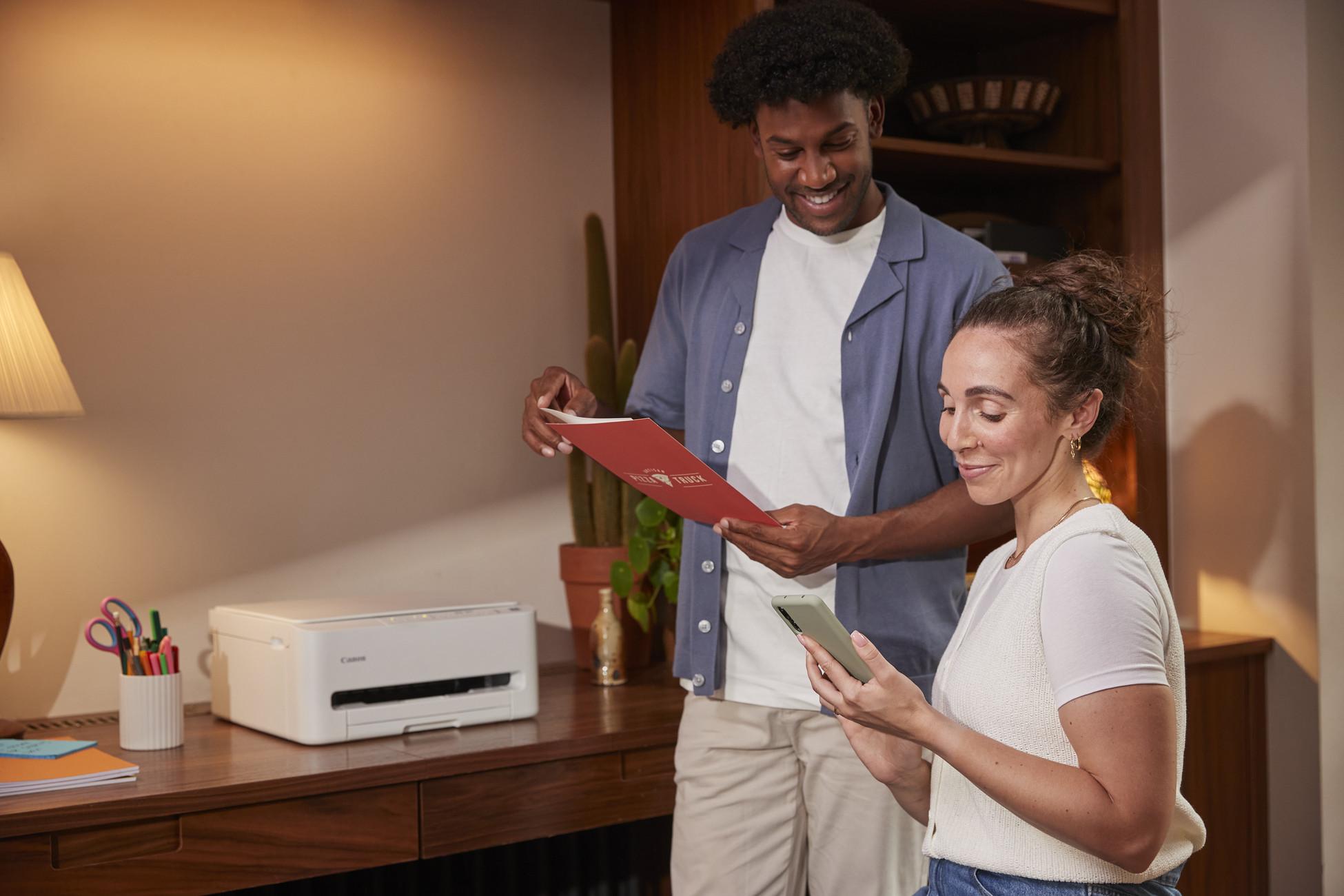 Mężczyzna i kobieta stoją przy biurku z drukarką, długopisami i książkami. Mężczyzna trzyma czerwony dokument.