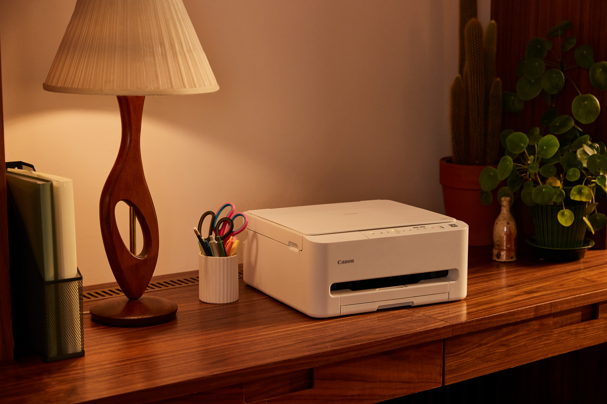 Biała drukarka Canon stoi na drewnianym biurku obok lampy i artykułów biurowych.