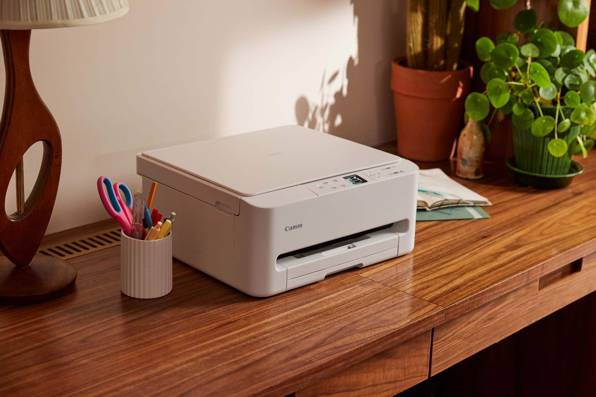 Biała drukarka Canon stoi na drewnianym biurku z lampą, roślinami i artykułami biurowymi.