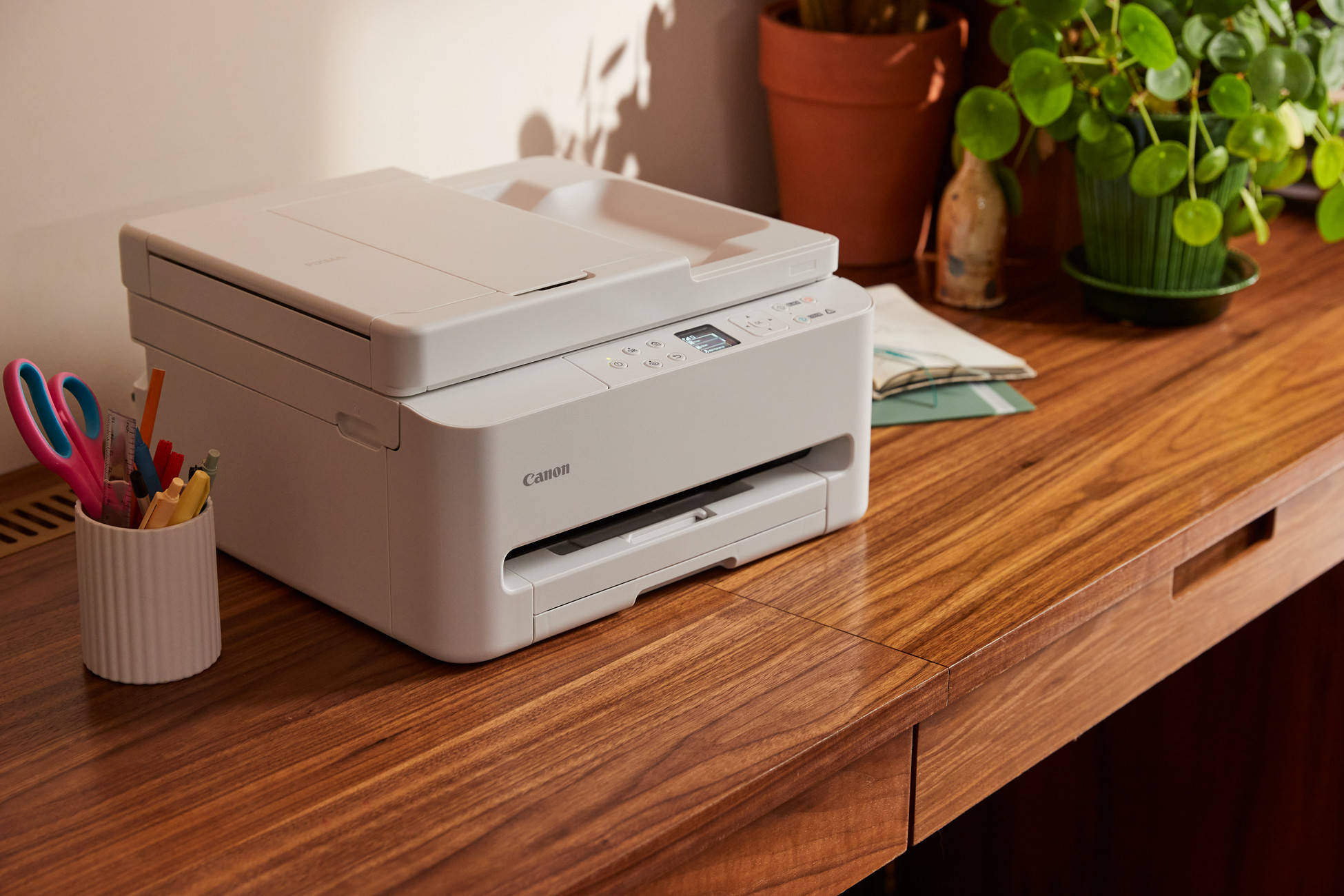 Biała drukarka Canon stoi na drewnianym biurku obok pojemnika na długopisy.