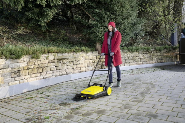 Kobieta w czerwonym płaszczu zamiata żółtą zamiatarką chodnikową podjazd.