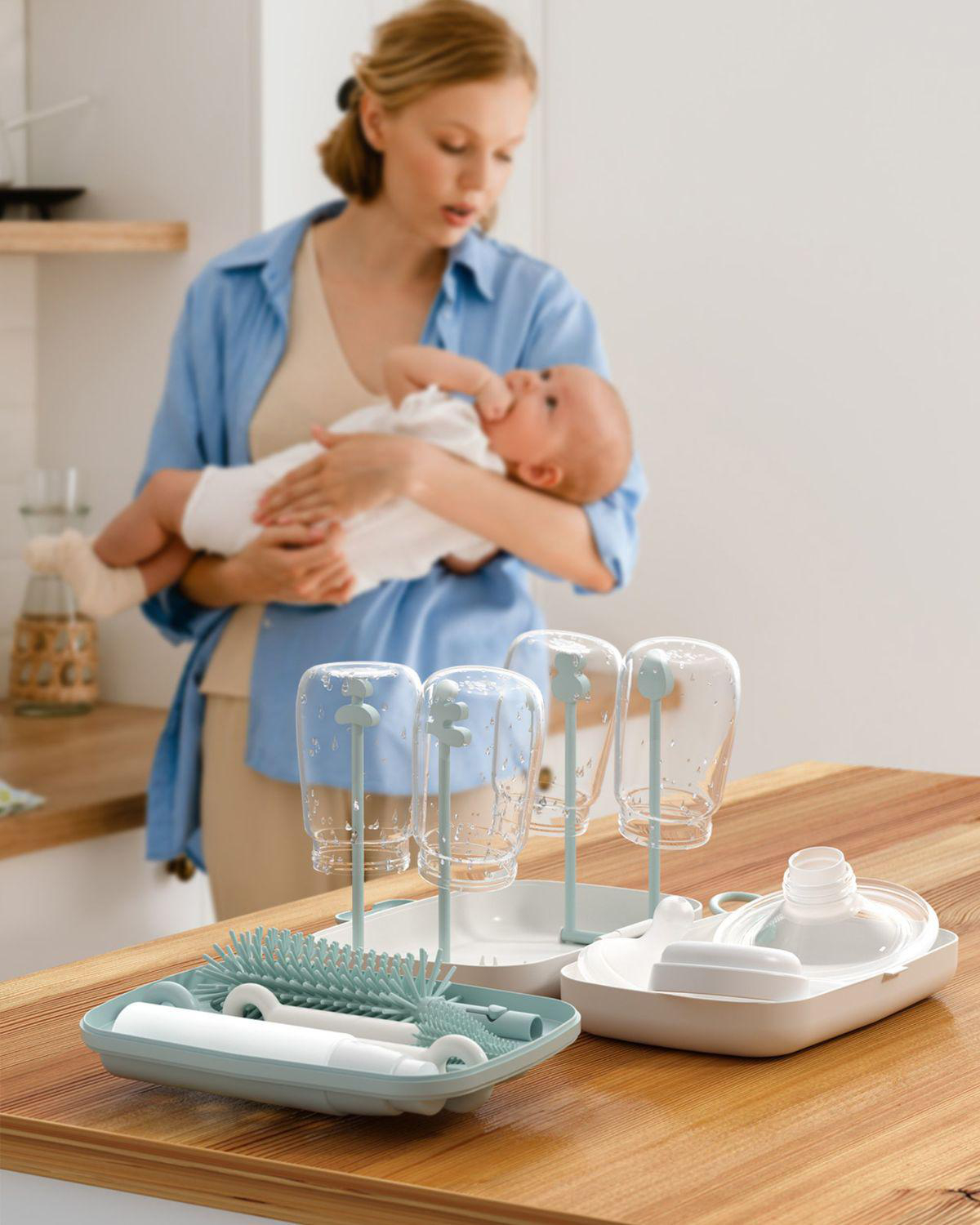 Frau hält Baby in der Nähe eines Trockengestells mit Flaschen und Bürsten auf einer Holztheke.